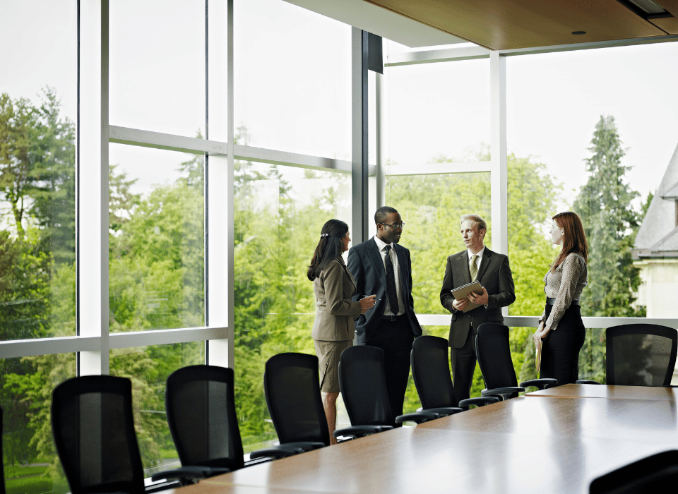Four business professionals talking in a bright conference room by large windows.