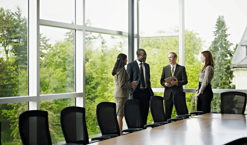 Four business professionals talking in a bright conference room by large windows.