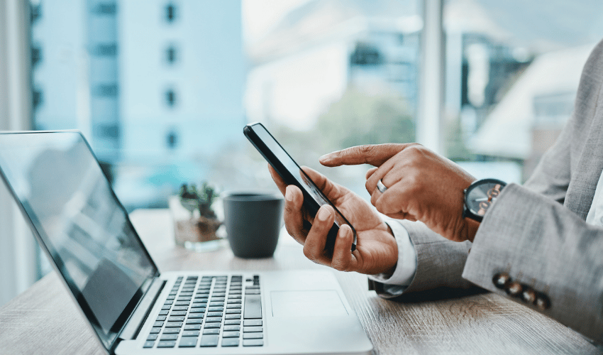 Person using smartphone beside laptop on desk in bright office setting